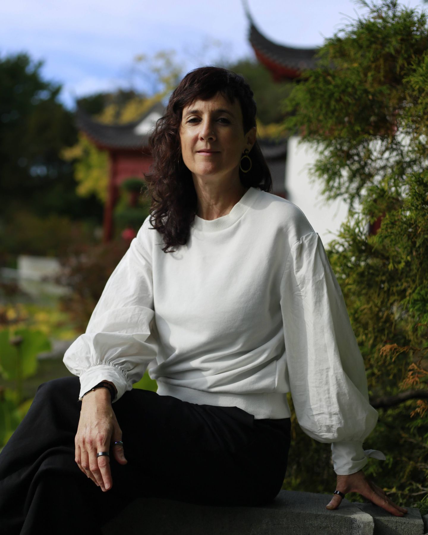 Julie Gazard, en blanc, assise sur un muret de pierre devant un pavillon du Jardin de Chine.